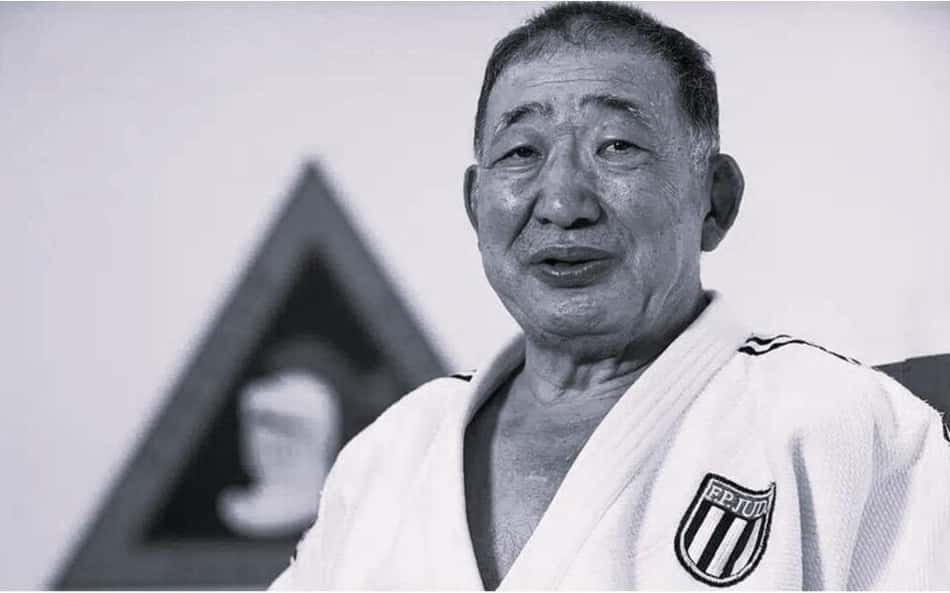 Judoca idoso em kimono branco, com emblema de competição no peito.