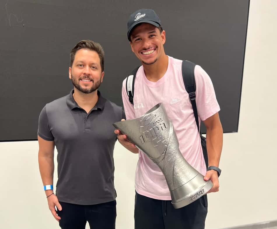 Dois homens posam com troféu prateado, sorrindo; um usa boné preto e camiseta rosa, o outro polo cinza.