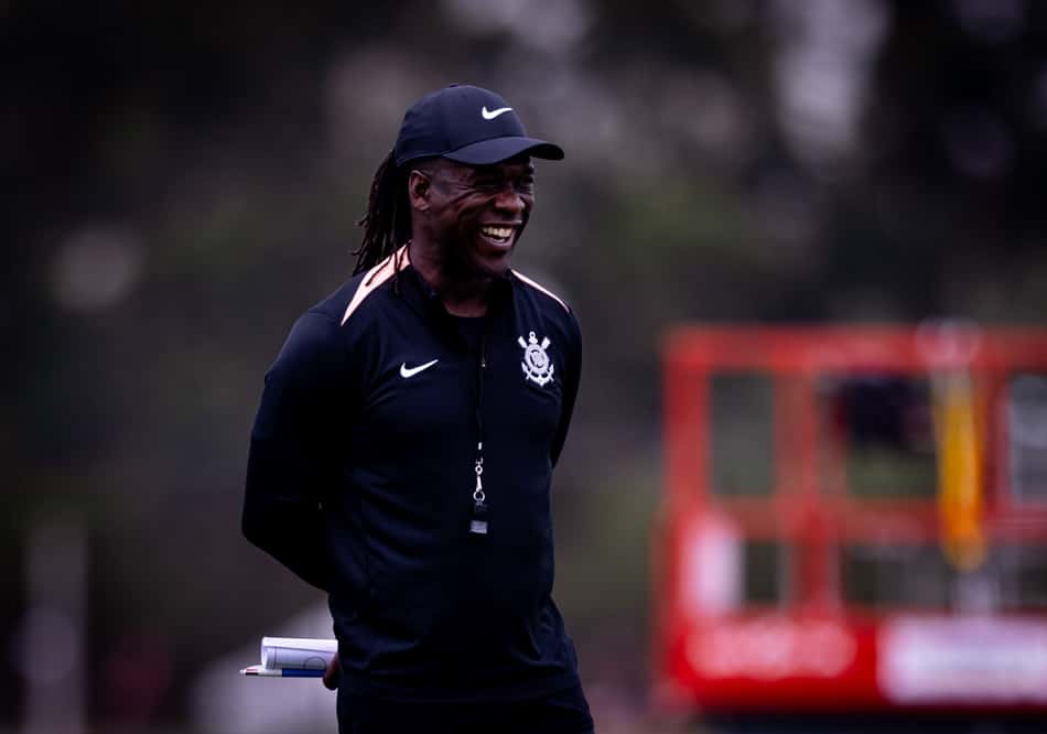 Clarence Seedorf sorrindo durante treino de futebol, com boné Nike e jaqueta preta.