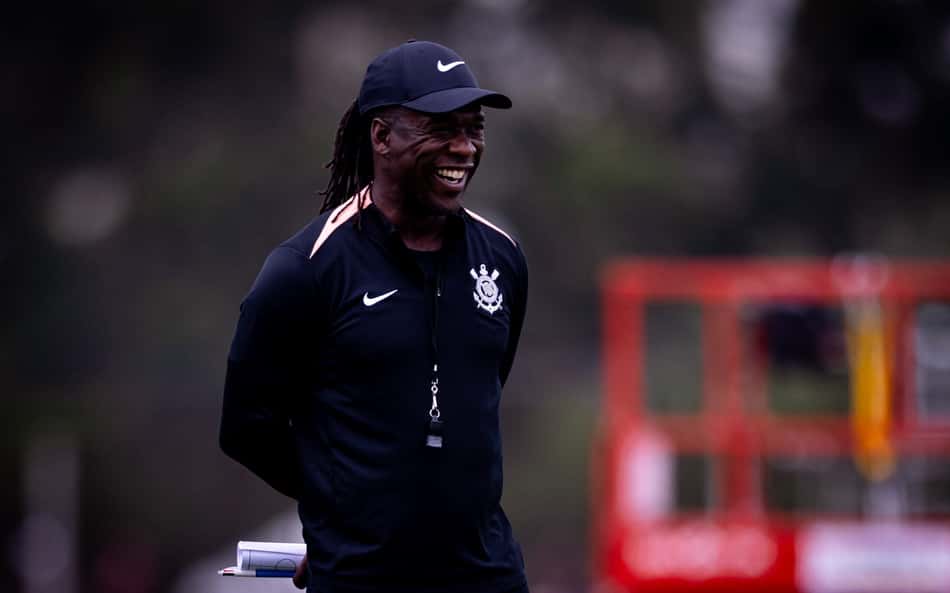 Clarence Seedorf sorrindo durante treino de futebol, com boné Nike e jaqueta preta.