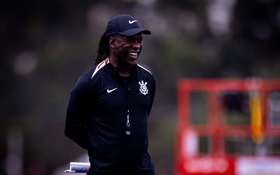 Clarence Seedorf sorrindo durante treino de futebol, com boné Nike e jaqueta preta.