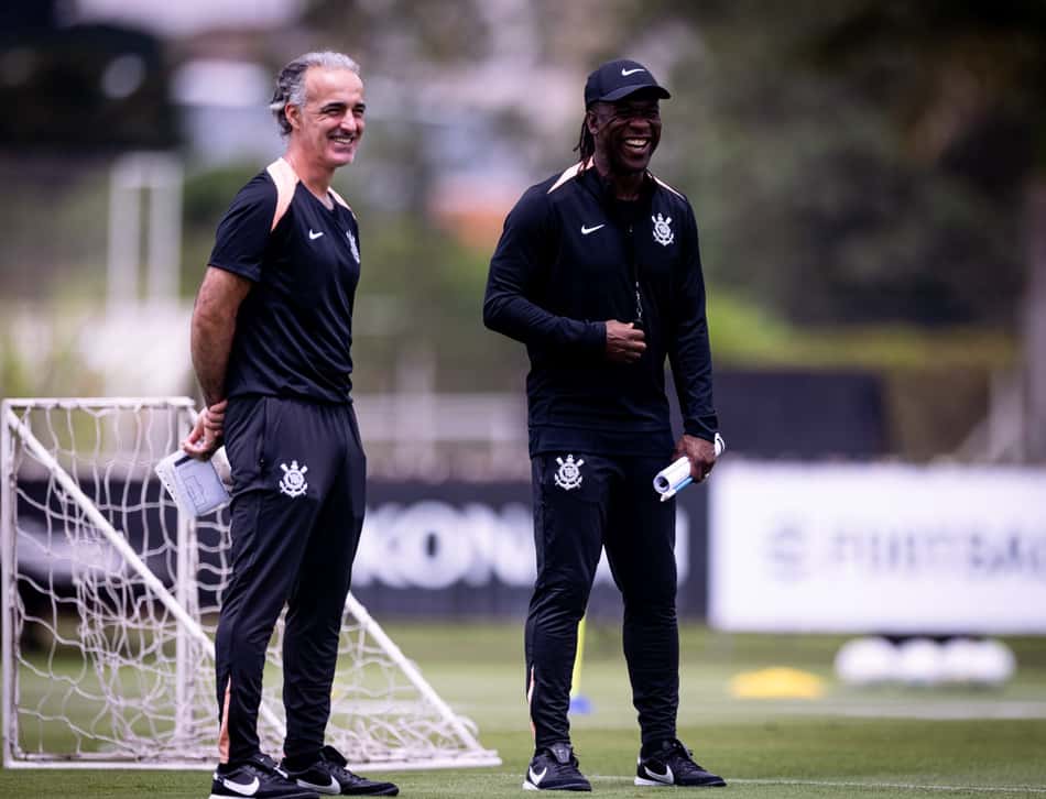 Dois membros da comissão técnica do Corinthians em treino de futebol, sorrindo no campo.