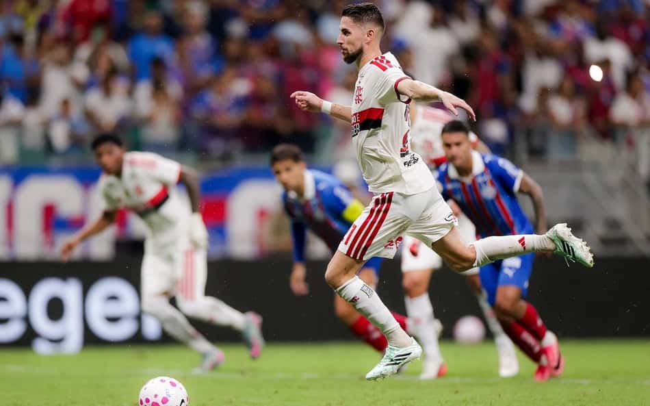 Jogador em camisa branca com listras vermelhas corre para cobrar pênalti; adversários azul e vermelho ao fundo.
