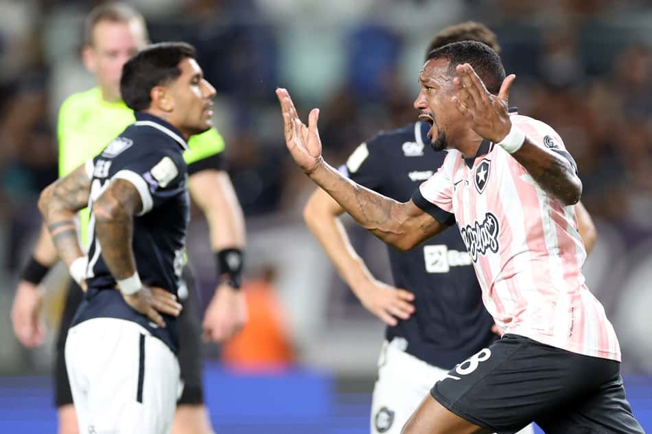Jogador com camisa rosa e branca levanta os braços, cercado por adversário em uniforme escuro.