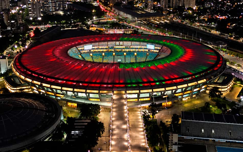 Estádio do Maracanã ao entardecer, arquibancadas iluminadas em vermelho e verde, cidade iluminada ao redor.