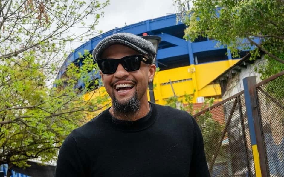 Felipe Melo com boné cinza, óculos escuros e casaco preto sorri ao ar livre, com grade e estrutura azul/amarela ao fundo