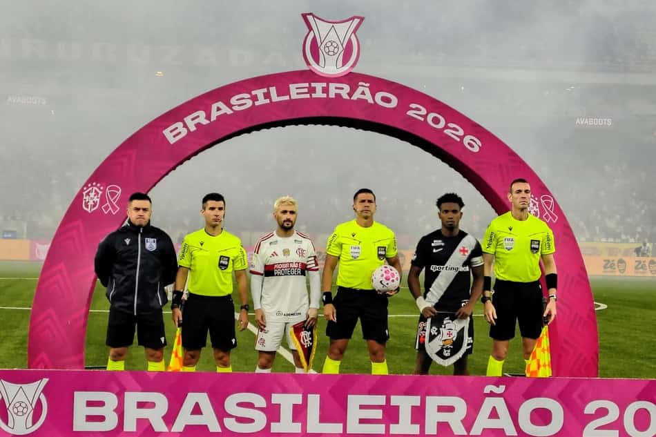 Seis indivíduos posam sob arco rosa com BRASILEIRÃO 2026; árbitros em amarelo e jogadores com uniformes variados.