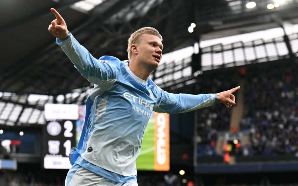 Jogador do Manchester City comemora gol com os braços abertos em estádio lotado.
