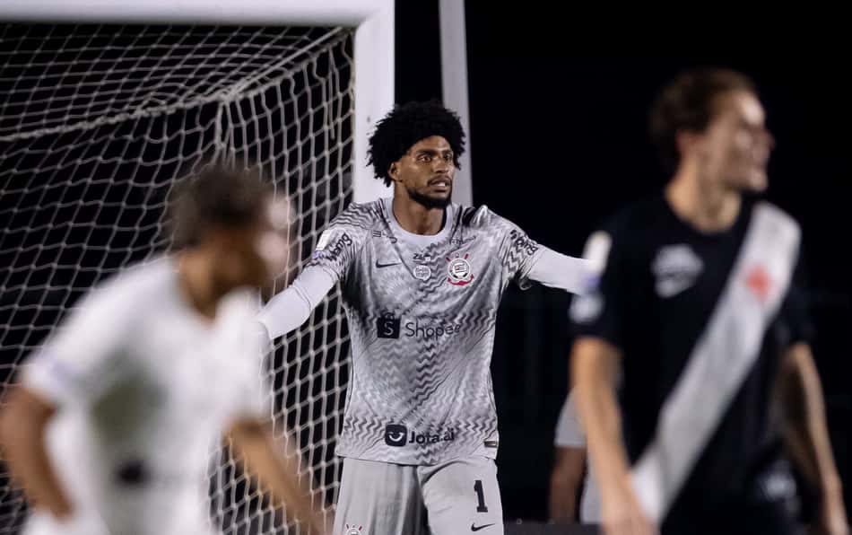 Goleiro em uniforme cinza, braços abertos diante do gol durante partida noturna.