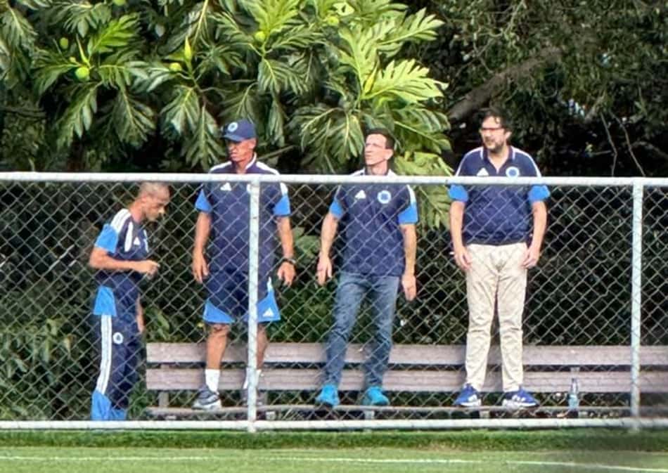 Quatro dirigentes do Cruzeiro em pé junto à grade, com banco ao fundo, num campo de futebol cercado por árvores.