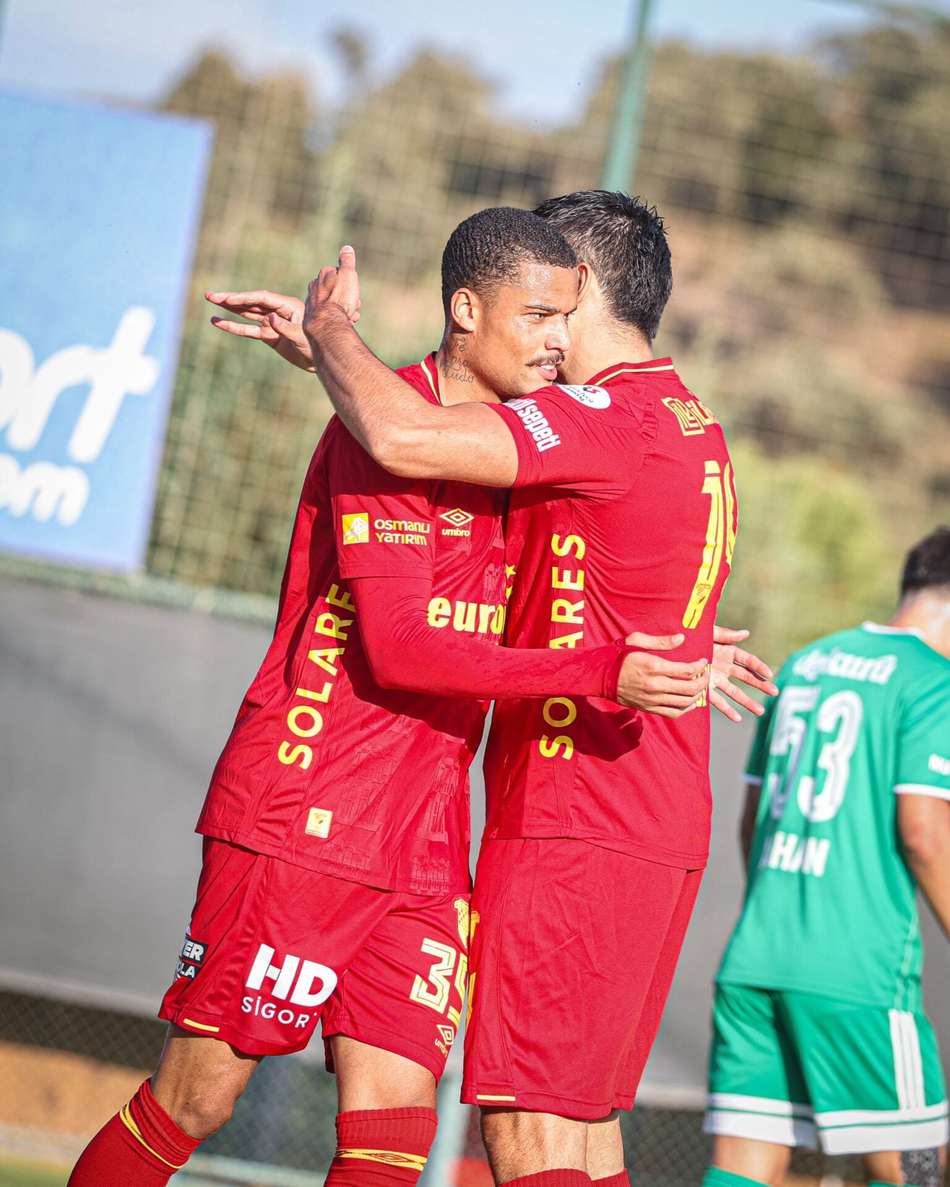 Janderson abraça colega de time vermelho em campo durante a partida, com jogador verde ao fundo