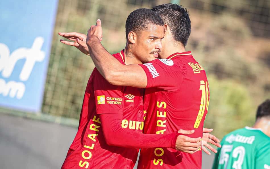 Janderson abraça colega de time vermelho em campo durante a partida, com jogador verde ao fundo
