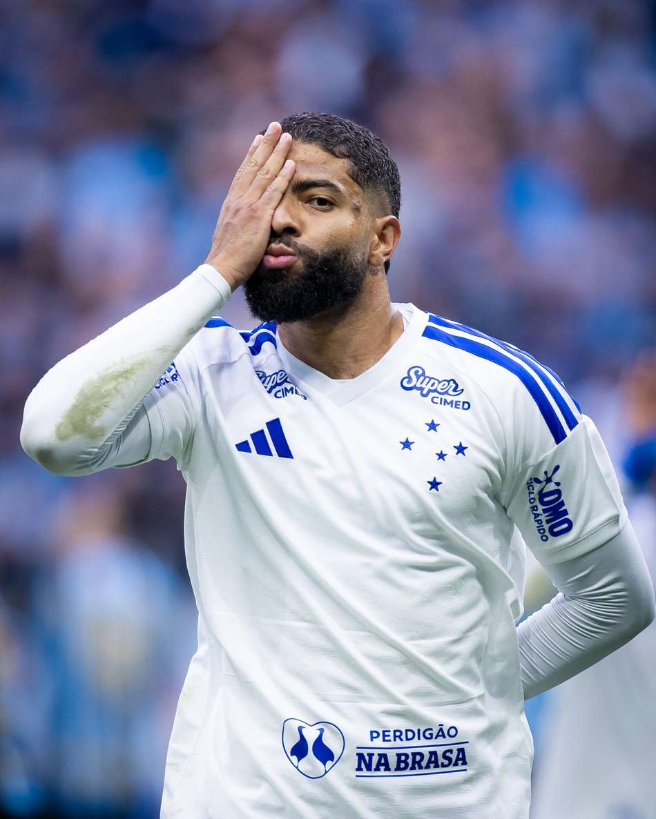 Jogador de futebol em uniforme branco com detalhes azuis, cobrindo o rosto com a mão em comemoração.