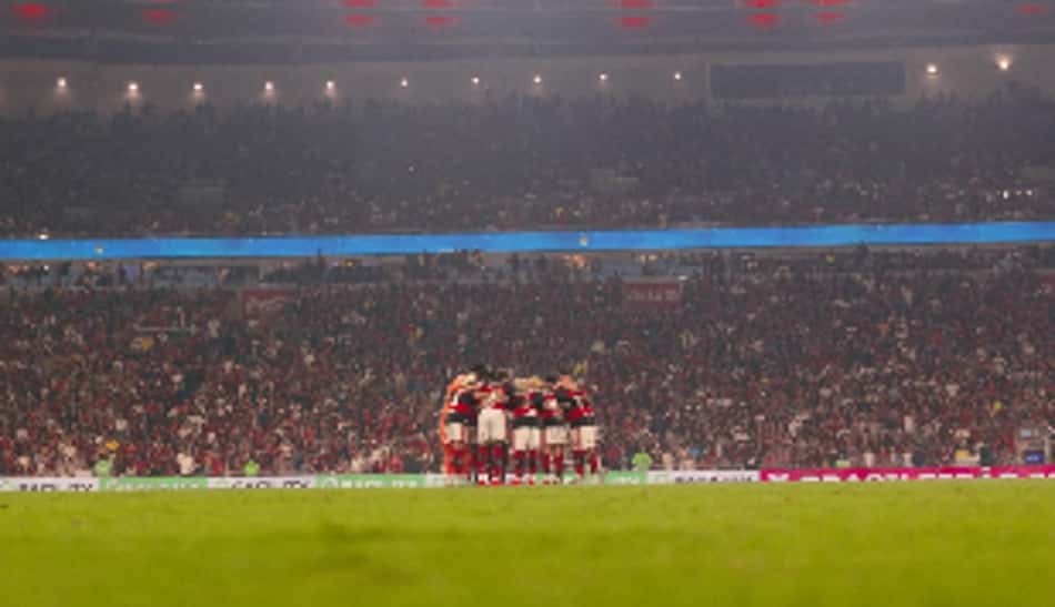 Jogadores do Flamengo formam um círculo no gramado, com torcida ao fundo.