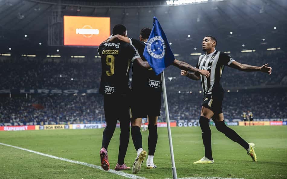 Trio de jogadores celebra gol no clássico, junto à bandeira de escanteio, diante da torcida.