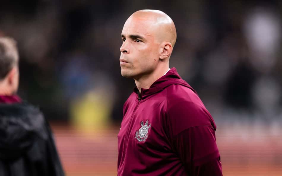 Júlio César, jogador com cabeça raspada, veste casaco vinho com emblema da equipe, em campo.