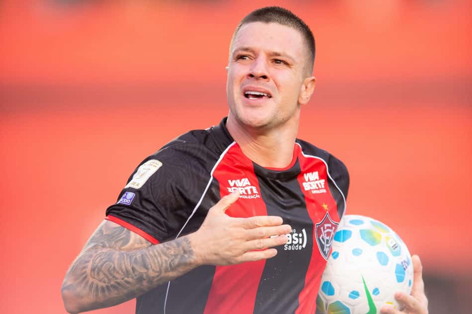 Jogador com uniforme listrado preto e vermelho celebra gol, segurando a bola com tatuagens visíveis no braço.