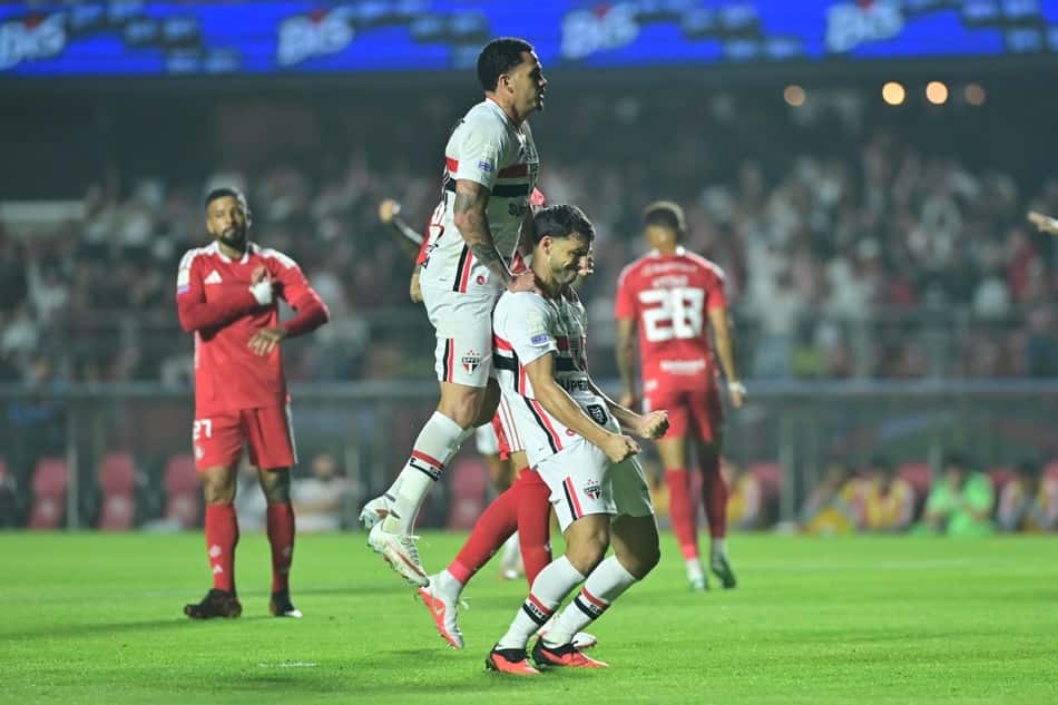 Dois jogadores do São Paulo celebram gol em campo, com torcedores ao fundo.