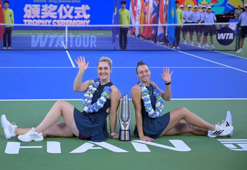 Luisa Stefani e parceira comemoram vitória de duplas, sentadas na quadra com troféu entre elas e colares de flores.