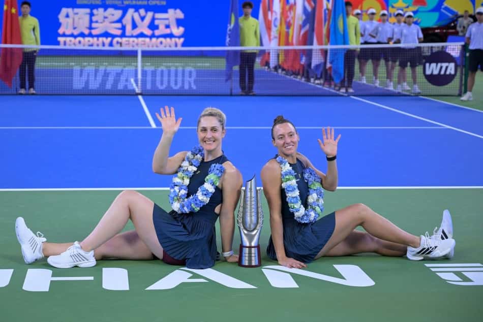 Luisa Stefani e parceira comemoram vitória de duplas, sentadas na quadra com troféu entre elas e colares de flores.