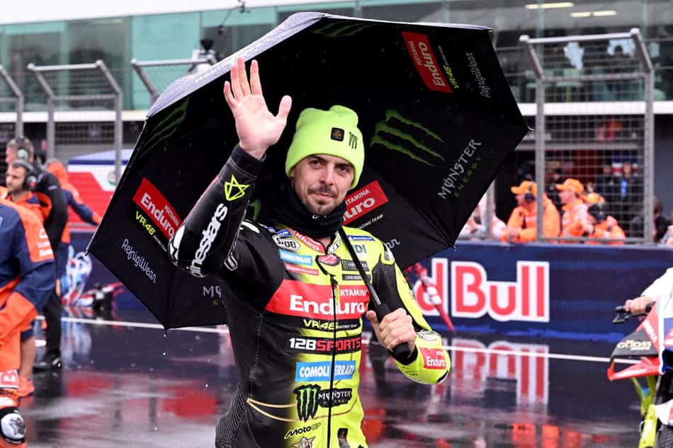 Piloto de MotoGP sob guarda-chuva, macacão preto e amarelo com patrocínios, acenando para a câmera.