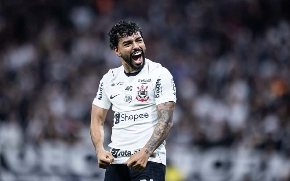 Jogador de futebol em camisa branca celebra em campo, com tatuagens nos braços, estádio lotado ao fundo.