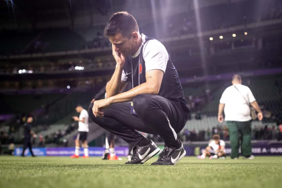 curopos agachado no gramado de estádio, vestindo camiseta preta com detalhes brancos, mão no queixo, ao fundo torcedores.