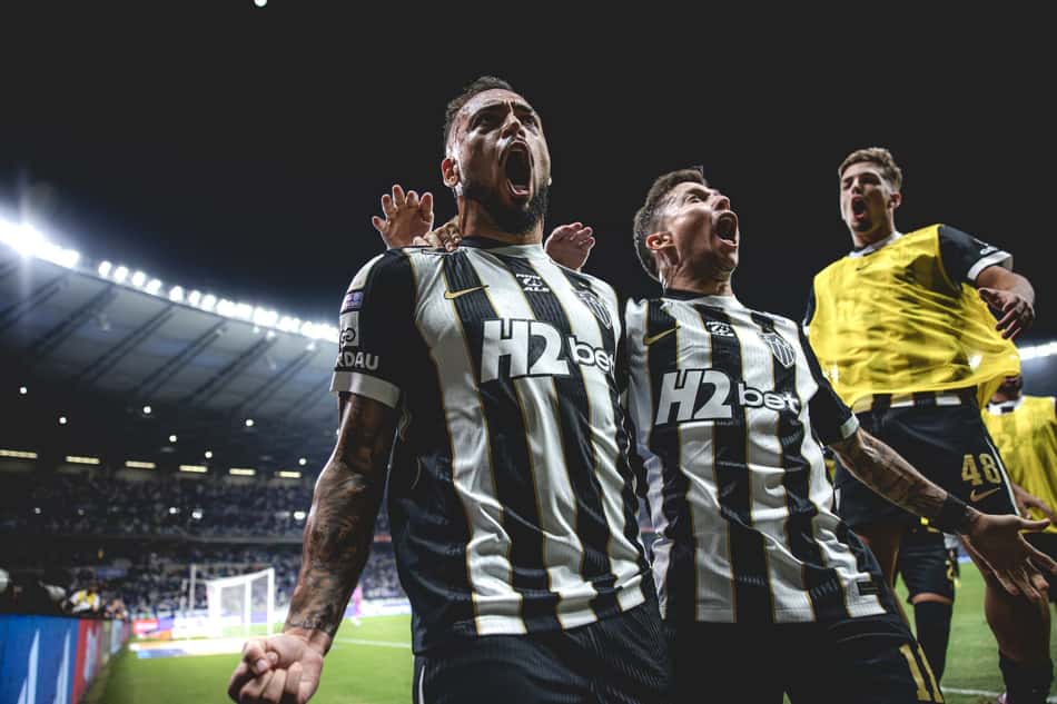 Maycon e Bernard celebram gol em jogo de futebol, torcedores ao fundo.