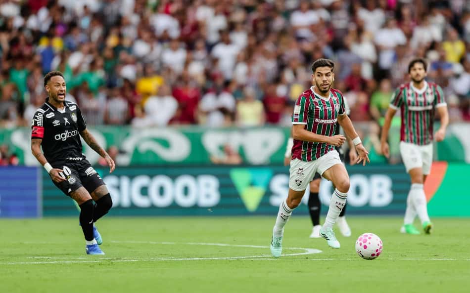 Jogador do Fluminense com a bola em ataque veloz, cercado por adversário com uniforme preto, estádio lotado ao fundo.