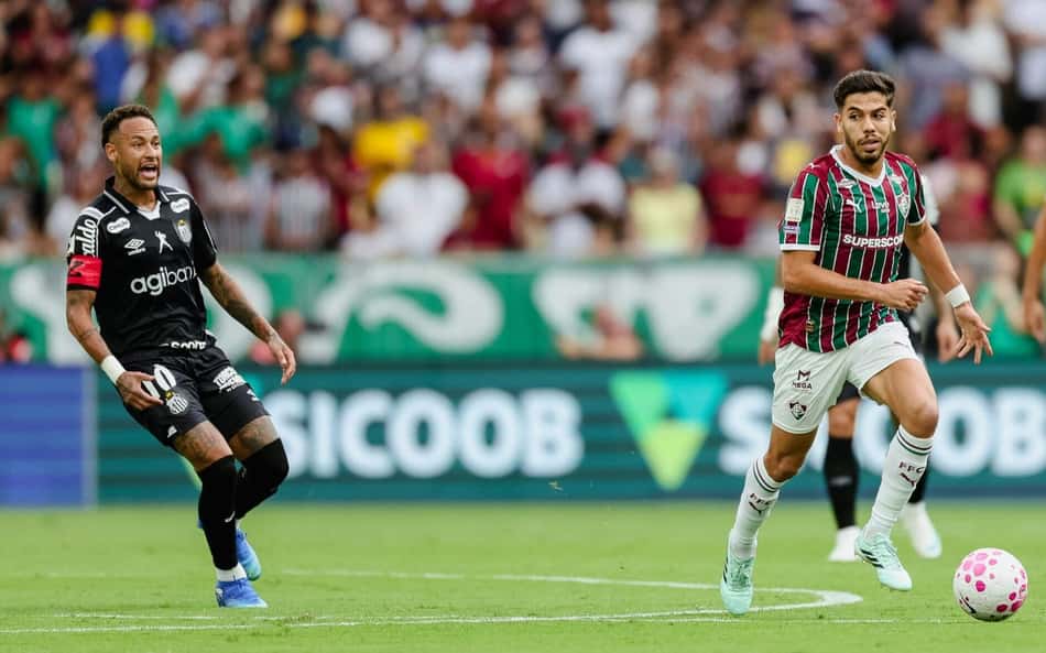 Jogador do Fluminense com a bola em ataque veloz, cercado por adversário com uniforme preto, estádio lotado ao fundo.