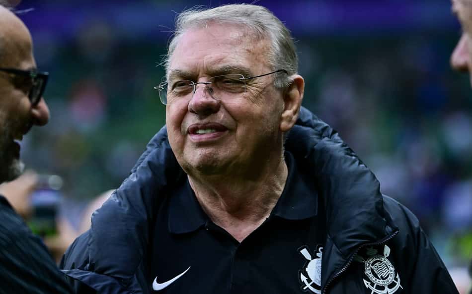 Homem idoso com óculos, casaco acolchoado e brasão no peito, sorrindo em estádio durante evento esportivo.