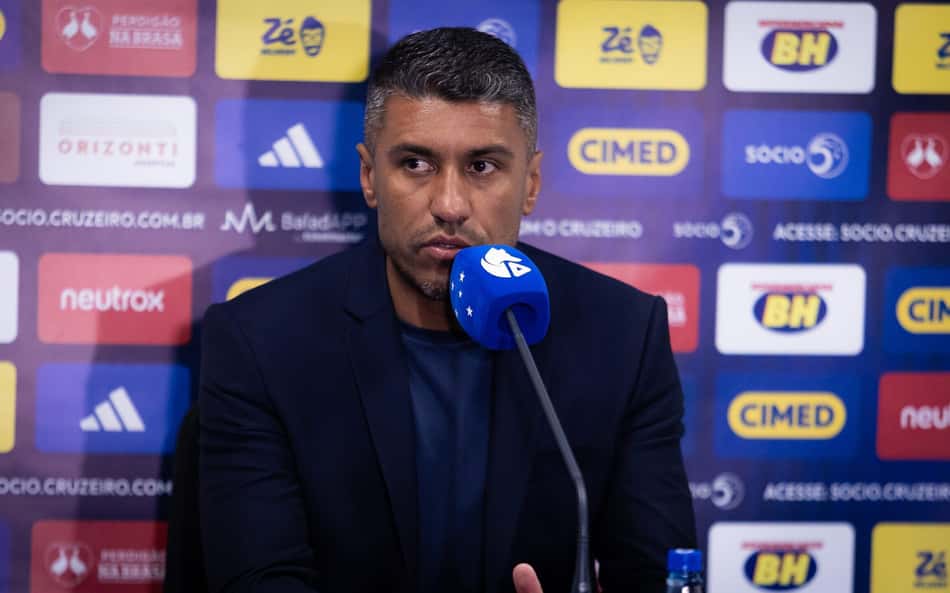 Homem em coletiva de imprensa com microfone azul, discutindo a arbitragem da partida Grêmio x Cruzeiro.Paulinho critica arbitragem de Grêmio x Cruzeiro