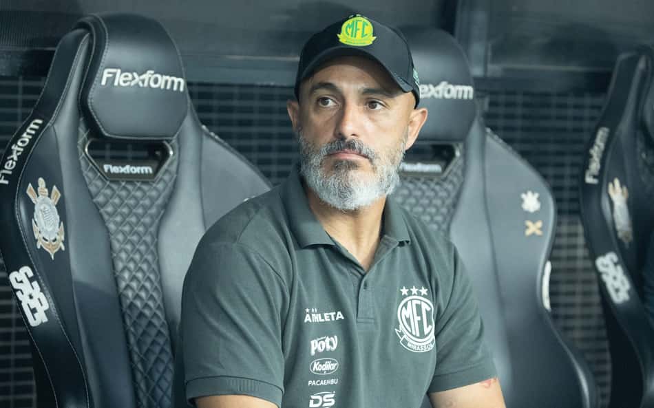 Treinador com polo verde e boné preto, sentado no banco de reservas de futebol.
