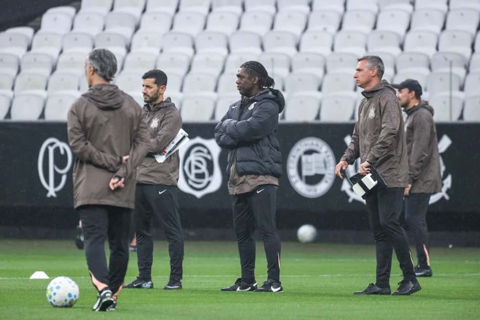 Grupo de treinadores do Corinthians em treino, casacos marrons, observando a prática no campo com bolas.