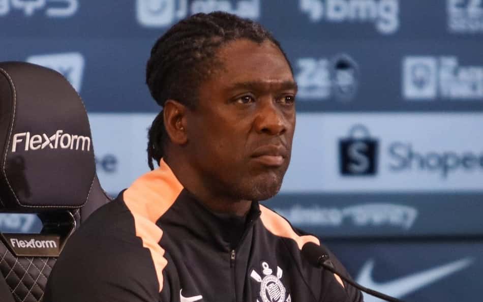 Clarence Seedorf em coletiva do Corinthians, jaqueta preta com escudo, diante de microfone.
