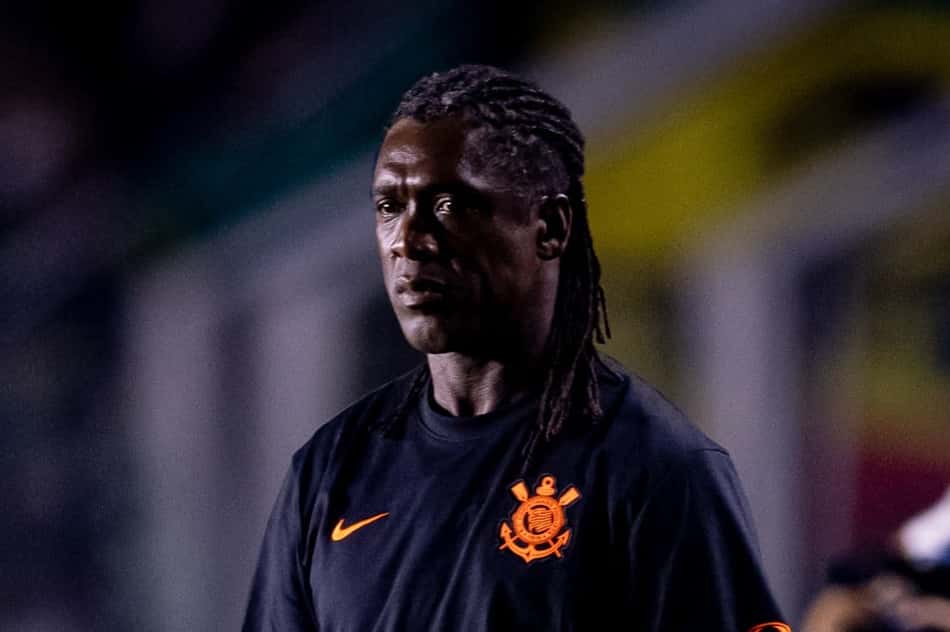 Seedorf, técnico, em uniforme preto com emblema laranja, em estádio ao fundo.