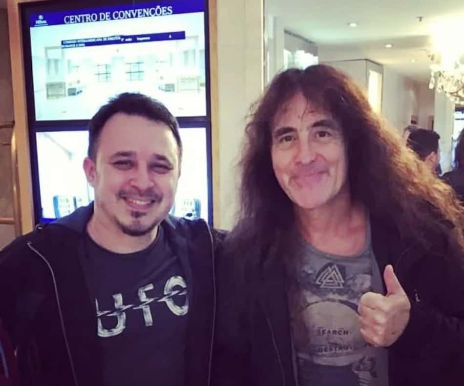 Steve Harris e Rodrigo sorrindo em centro de convenções; Rodrigo faz sinal de positivo.