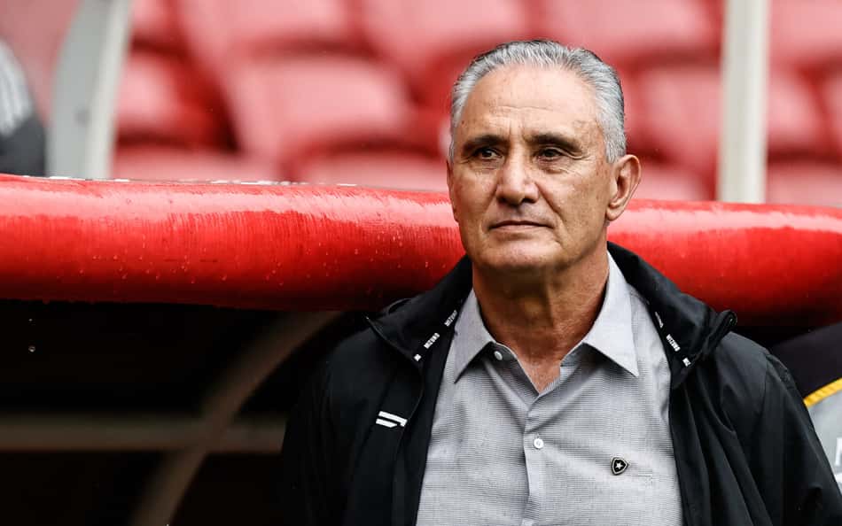 Tite, técnico do Botafogo, em área técnica, com camisa cinza e jaqueta preta, diante de arquibancadas vermelhas.