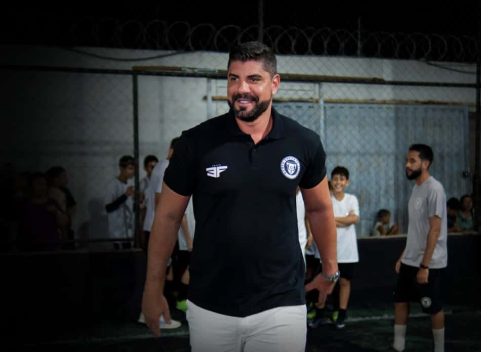 Victor Oliveira North, treinador com polo preto, logotipo no peito, em treino com jovens ao fundo.