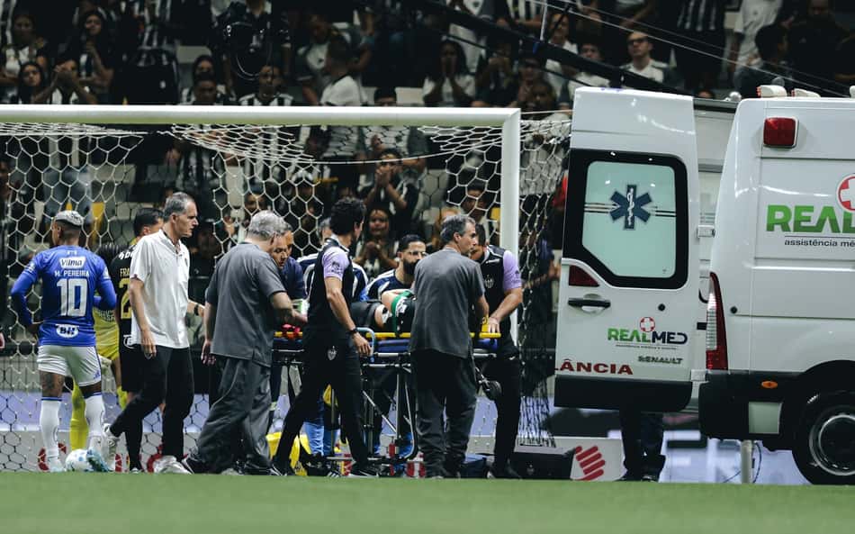 Jogador com dorsal 10 sai de ambulância cercado pela equipe médica durante jogo de futebol; torcedores ao fundo.