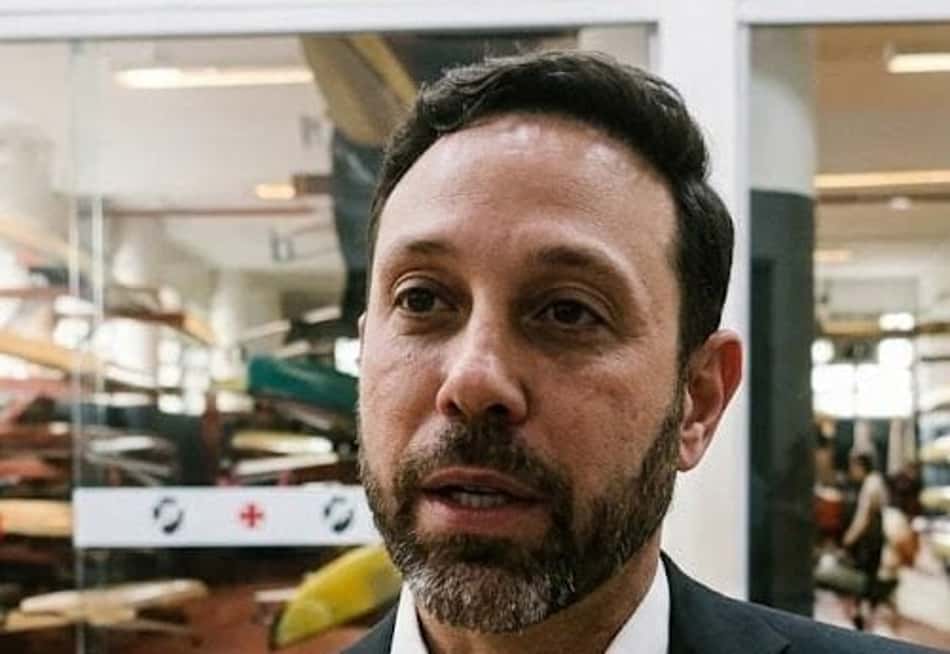 Alan Vasco, em entrevista, blazer preto, com microfone; fundo com pranchas de surf armazenadas.
