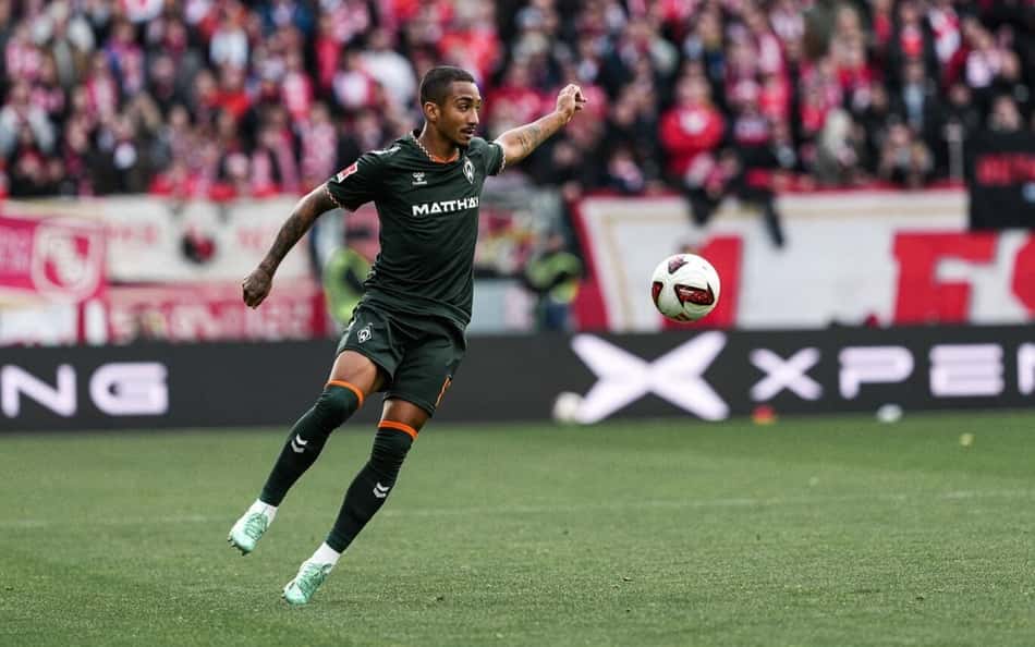 Jogador do Werder Bremen domina a bola em campo, com torcida ao fundo.