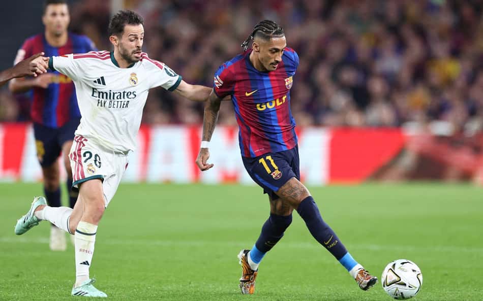 Jogador do Barcelona em uniforme azul e grená dribla a bola enquanto jogador do Real Madrid acompanha a jogada.