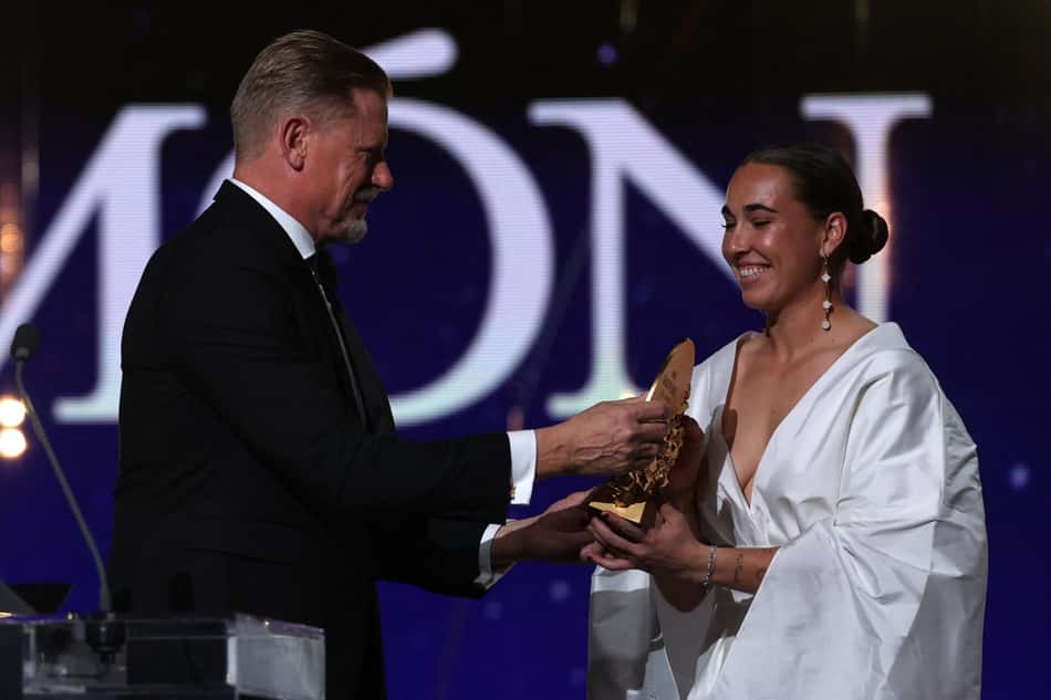 Homem de terno entrega troféu à mulher de vestido branco em palco, com letras grandes ao fundo.