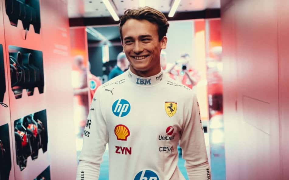 Dino Beganovic, piloto Ferrari na F1, sorrindo no corredor com macacão branco e logos da equipe.