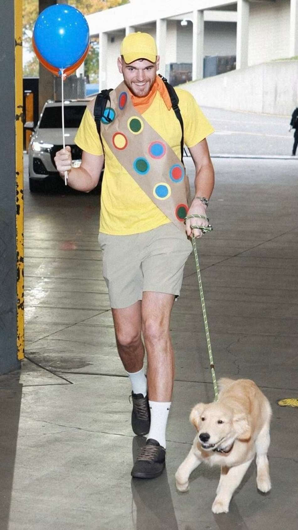 Homem com camiseta amarela, boné amarelo, faixa com círculos coloridos, segurando balão azul, passeando com cachorro Golden Retriever