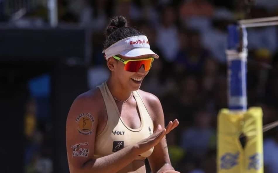 Jogadora de vôlei de praia sorri na quadra, com viseira branca Red Bull e shorts pretos