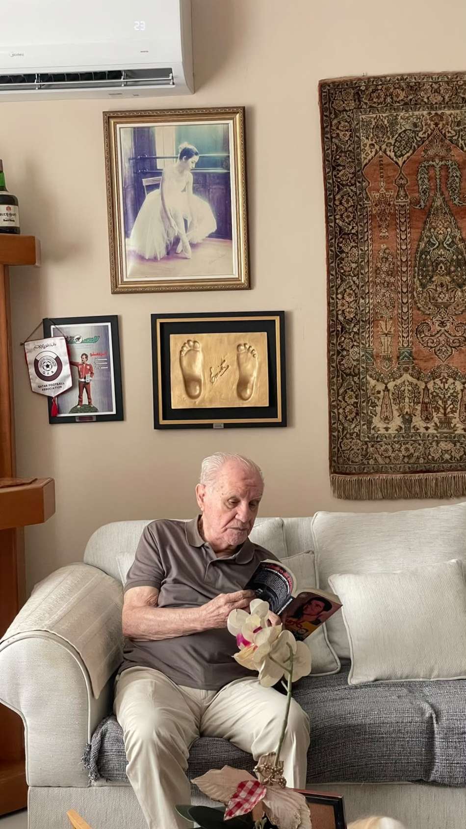 Evaristo Macedo lendo livro no sofá, em sala com quadros na parede e tapeçaria ao fundo.