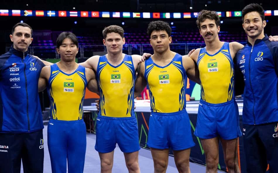 Seis ginastas brasileiros em amarelo e azul, com dois técnicos, posam em arena no Mundial de ginástica artística.