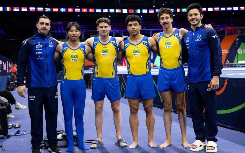 Seis ginastas brasileiros em amarelo e azul, com dois técnicos, posam em arena no Mundial de ginástica artística.